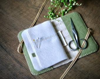 May include: A white felt sewing kit with a green felt border. The kit has a small pocket for pins, a compartment for scissors, and a small ruler. The kit is closed and has a ribbon tied around it.