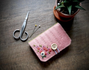 May include: A pink felt needle case with embroidered flowers and a button closure. Silver scissors and two pins are next to the case on a wooden surface. A small potted succulent plant is in the background.