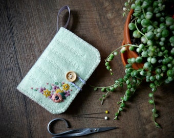 May include: A light green felt pouch with a floral embroidery design and a wooden button closure. The pouch is sitting on a wooden surface with a potted plant in the background.