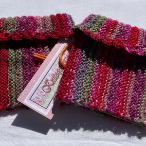 May include: Two pairs of knitted wrist warmers in a multi-colored striped pattern of red, pink, green, and purple. The wrist warmers have a crocheted edge and are displayed on a white surface with a pink and green tag that says "A Stitch in Time".