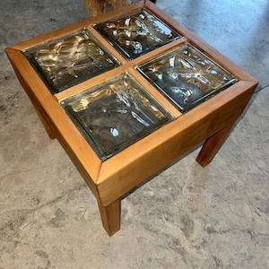 May include: A square coffee table made of light brown wood, featuring four glass block inserts. The table has four legs and a wooden frame. The glass blocks are clear and textured, creating a distinctive design.