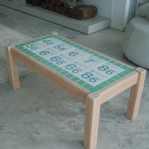 May include: A rectangular wooden coffee table with a tiled top. The tiles are white with blue numbers and green borders. The table's legs are light-coloured wood. The numbers on the tiles include 46, 56, 6, 7, and 86.