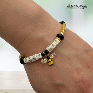 May include: A beaded bracelet with white lettered cubes spelling "BUSY BEE" and a gold-colored bee charm with rhinestone accents. The bracelet has black and gold beads and the text "Ticket to Magic".