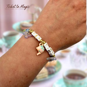 May include: A beaded bracelet featuring white square letter beads spelling TEA TIME, interspersed with metallic gold spherical beads. The bracelet includes a small peach-colored teapot charm with floral accents and a translucent purple butterfly charm.