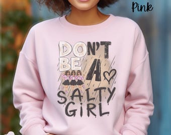 May include: A light pink crewneck sweatshirt featuring a graphic print with the text "DON'T BE A SALTY GIRL" in black and beige lettering. The design includes an illustration of three figures in pink jackets and black skirts, a heart, and rain streaks.