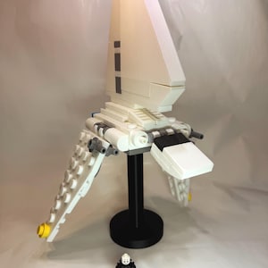 May include: A white and gray model spaceship with a black stand and a small figurine. The spaceship has angled wings and a tall, triangular body. Yellow accents are visible on the wings and top. The figurine is gray and black.