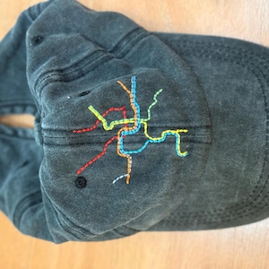 May include: A dark gray baseball cap with a colorful embroidered map design. The map features lines in red, yellow, blue, and green. The cap has a curved brim and a black button on top.