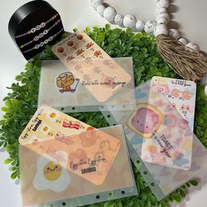 May include: A collection of clear, plastic budget envelopes with colorful, food-themed designs. The envelopes feature illustrations of eggs, bacon, toast, and cinnamon rolls, with text like "Egg-stra Egg-stra Savings" and "Roll into sweet savings."