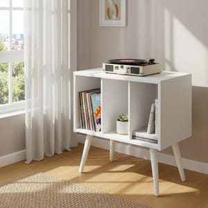 May include: A white storage cabinet with three open compartments supported by four tapered legs. The left compartment holds vinyl records, the center holds a small potted succulent, and the right holds books. A portable record player sits on top.