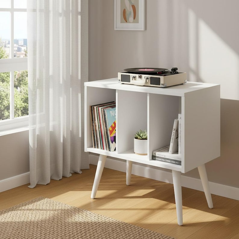 May include: A white storage cabinet with three open compartments supported by four tapered legs. The left compartment holds vinyl records, the center holds a small potted succulent, and the right holds books. A portable record player sits on top.