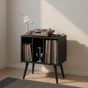 May include: A black wooden storage cabinet with four tapered legs, featuring three open compartments filled with vinyl records and a pair of headphones. A wooden record player, a glass vase with white flowers, and books sit on top.