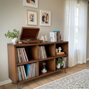Pode incluir: Um toca-discos de madeira está em cima de uma unidade de armazenamento de seis cubos marrom escuro, cheia de discos de vinil. A unidade tem pernas de grampo de metal e é decorada com plantas, livros e objetos decorativos. Obras de arte penduradas na parede acima.