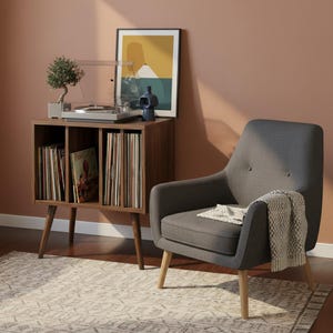 May include: A dark gray upholstered armchair with light wood legs, featuring a textured throw blanket. Beside it, a wooden record storage cabinet holds vinyl records, a turntable, a small bonsai tree, and a blue vase, with framed art above.