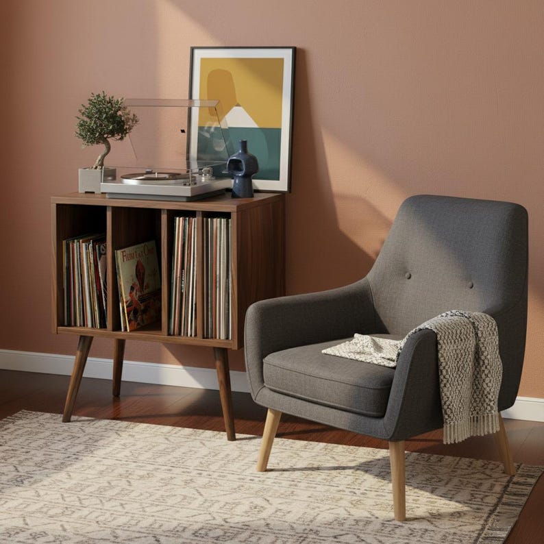 May include: A dark gray upholstered armchair with light wood legs, featuring a textured throw blanket. Beside it, a wooden record storage cabinet holds vinyl records, a turntable, a small bonsai tree, and a blue vase, with framed art above.