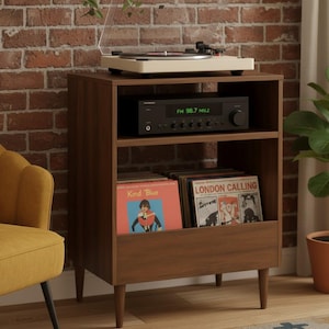 Puede incluir: Un mueble de madera marrón para tocadiscos con un tocadiscos color crema en la parte superior. El mueble tiene tres estantes, uno con un receptor estéreo que muestra "FM 98.7 MHZ". Los discos de vinilo se guardan en el estante inferior.