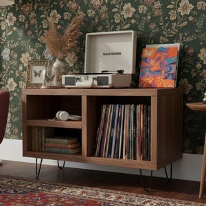 May include: A rectangular wooden media console with four black metal hairpin legs. The unit features an open shelf on the left holding white headphones and stacked books, and a large open compartment on the right filled with vinyl records.