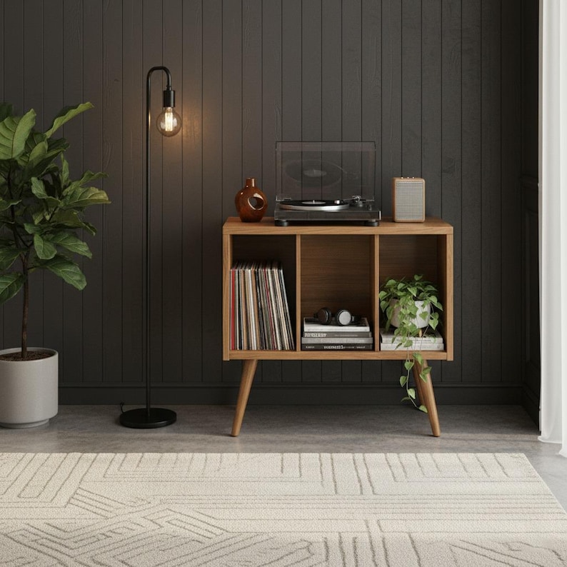 May include: A wooden record player console with three open storage compartments, featuring a turntable on top, a collection of vinyl records, books, and a small potted plant. A tall black floor lamp stands to the left of the wooden cabinet.