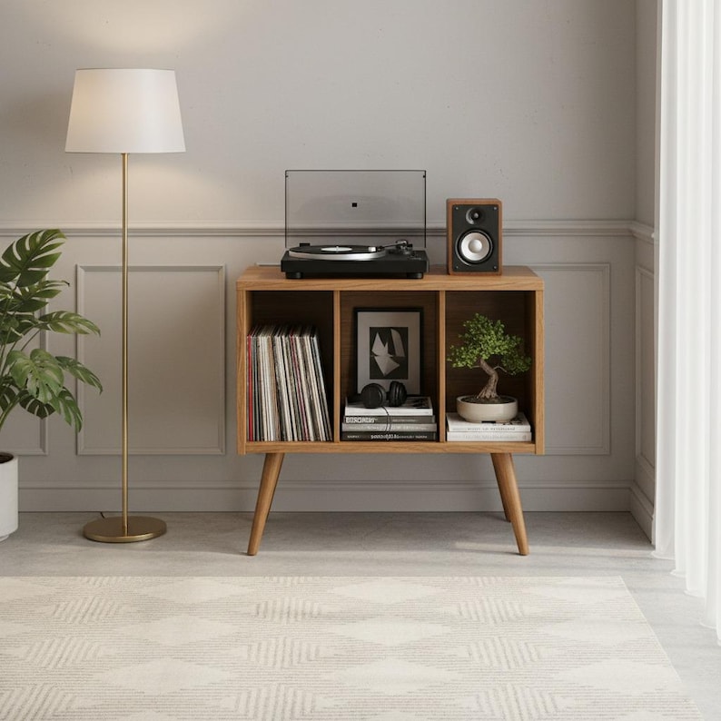 May include: A wooden media console with three open storage compartments, supported by four tapered legs. The top surface holds a black turntable and a wooden speaker. The shelves contain vinyl records, a framed print, books, and a small bonsai tree.