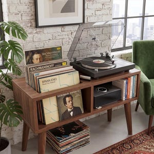 May include: A walnut-finished wooden media console with tapered legs, featuring a black turntable with a clear dust cover on top, a built-in storage compartment for vinyl records, a lower shelf for audio equipment, and a stack of records below.