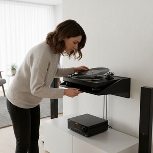 K&ouml;nnte beinhalten: Ein schwarzer Plattenspieler an einem Wandregal, mit einer Schallplatte auf dem Teller. Darunter steht ein schwarzer Stereo-Receiver auf einem wei&szlig;en Schrank. Eine Frau legt eine Schallplatte auf den Plattenspieler.