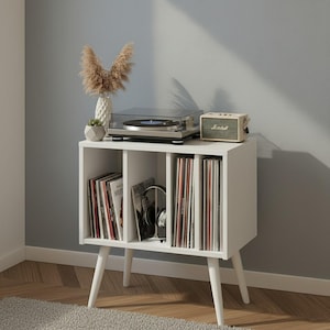 May include: A white wooden storage cabinet with four vertical compartments for vinyl records, supported by four tapered legs. On top sits a black turntable, a beige speaker with the Marshall logo, and a white vase containing dried pampas grass.