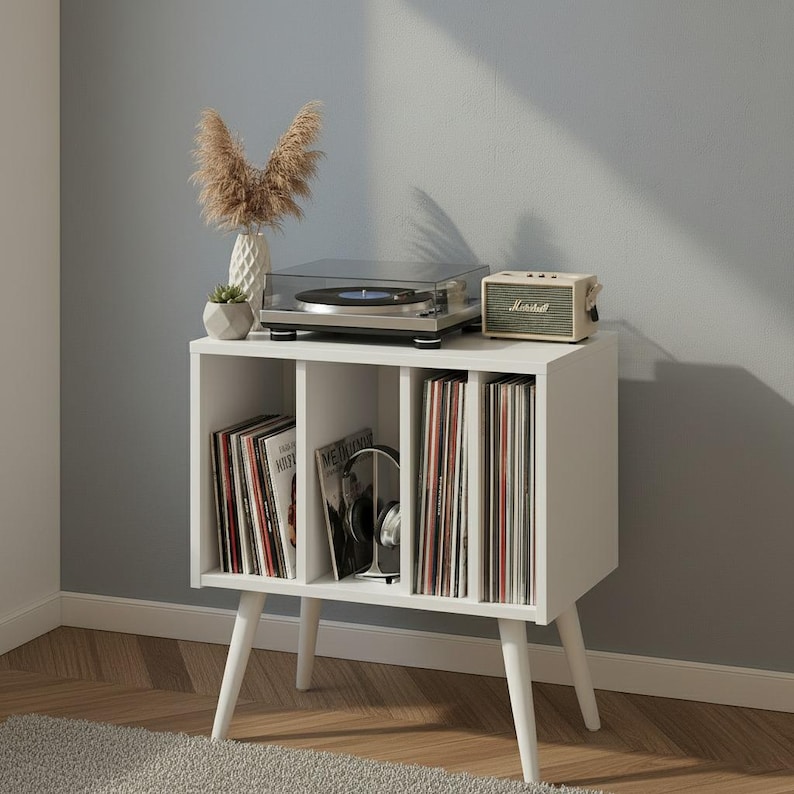May include: A white wooden storage cabinet with four vertical compartments for vinyl records, supported by four tapered legs. On top sits a black turntable, a beige speaker with the Marshall logo, and a white vase containing dried pampas grass.