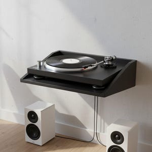 K&ouml;nnte beinhalten: Schwarzer, an der Wand montierter Plattenspieler mit einer Vinyl-Schallplatte. Der Plattenspieler steht auf einem schwarzen Regal, darunter zwei wei&szlig;e Lautsprecher. Das Setup befindet sich vor einer wei&szlig;en Wand und wirkt minimalistisch.
