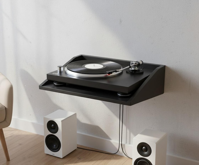 K&ouml;nnte beinhalten: Schwarzer, an der Wand montierter Plattenspieler mit einer Vinyl-Schallplatte. Der Plattenspieler steht auf einem schwarzen Regal, darunter zwei wei&szlig;e Lautsprecher. Das Setup befindet sich vor einer wei&szlig;en Wand und wirkt minimalistisch.