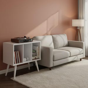 May include: A white wooden storage unit with three open compartments, supported by four tapered legs. The top surface holds a dark brown record player. Inside the compartments are vinyl records, a small potted plant, books, and white headphones.