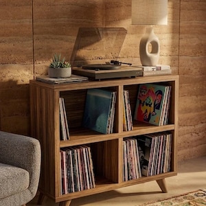 Peut inclure: Un meuble de rangement en bois pour disques vinyles doté de quatre compartiments ouverts remplis de disques. Sur le dessus se trouvent une platine noire avec un capot transparent, une petite plante succulente et une lampe beige.