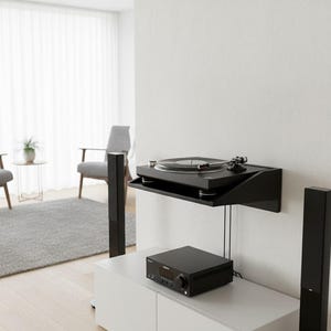 K&ouml;nnte beinhalten: Ein schwarzer Plattenspieler auf einem schwarzen Wandregal, darunter ein schwarzer Receiver. Zwei schwarze Lautsprecher flankieren einen wei&szlig;en Schrank. Der Raum hat ein minimalistisches Design mit grauen St&uuml;hlen und einem Teppich.