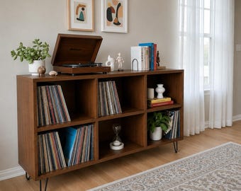 Puede incluir: Mueble de almacenamiento de discos de vinilo de estilo moderno. El mueble de madera marrón tiene seis compartimentos llenos de discos. Un tocadiscos se encuentra en la parte superior, junto con objetos decorativos y una pequeña planta. Tiene patas de horquilla de metal negro.