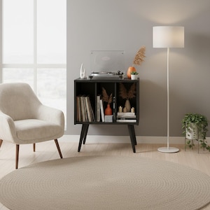 May include: A black wooden record player stand with three open storage compartments, supported by four tapered legs. The top surface holds a record player, while the shelves contain vinyl records, books, decorative vases, and small candles.