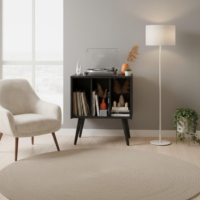 May include: A black wooden record player stand with three open storage compartments, supported by four tapered legs. The top surface holds a record player, while the shelves contain vinyl records, books, decorative vases, and small candles.