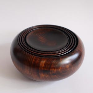 May include: A dark brown, polished wooden bowl with a smooth, rounded exterior and a series of concentric circular indentations on the top surface, creating a nested appearance within the center of the circular vessel.