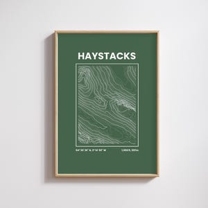 May include: Framed print with a dark green background, displaying a white topographic map of Haystacks. The word "HAYSTACKS" is at the top, with coordinates and elevation (597m) at the bottom. The frame is a light wood colour.