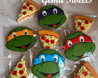May include: Cookies shaped like Teenage Mutant Ninja Turtles and pizza slices, individually wrapped in clear plastic. The cookies feature green turtle faces with different colored masks and pizza slices with red pepperoni.