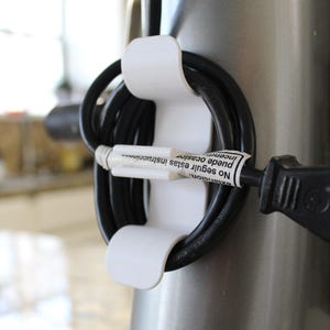 May include: A white cord organizer attached to a silver appliance, holding a black electrical cord. The cord is coiled and secured by the organizer. A label on the cord reads "No seguir estas instrucciones".