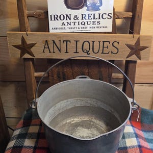 May include: A large, antique metal pot with a handle, sitting on a plaid blanket. Above it, a wooden sign with the word "ANTIQUES" and star accents. A sign for "Iron & Relics Antiques" is also visible.