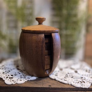 May include: A wooden coaster holder with a lid. The holder is a dark brown color with a lighter brown lid and a small knob on top. The holder has a slot on the side to hold the coasters. The coasters are visible in the slot.