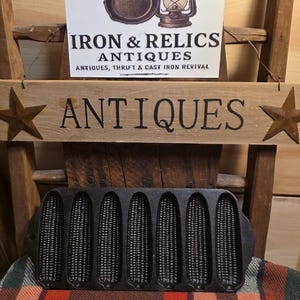 May include: A black cast iron corn stick pan with six molds, resting on a plaid blanket. Above, a sign reads "ANTIQUES" with star accents. A sign for "IRON & RELICS ANTIQUES" is also visible.