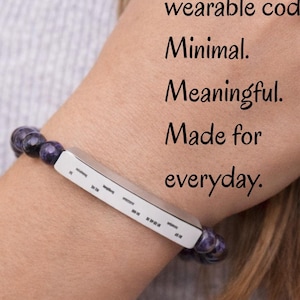 May include: A beaded bracelet with purple beads and a silver bar. The bar has a series of dashes and dots, with the text "AuDHD - in a wearable code. Minimal. Meaningful. Made for everyday."