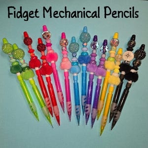 May include: A collection of Fidget Mechanical Pencils in various colors including green, red, pink, blue, purple, yellow, and black. Each pencil features decorative beads, small pom-poms, and textured elements on the top end.