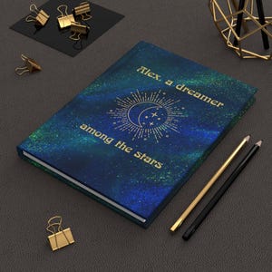 May include: A hardcover journal with a dark blue and green galaxy-themed cover featuring a gold sun and moon graphic. The text reads, "Alex, a dreamer among the stars." The journal is accompanied by a gold pencil, a black pencil, and several gold metal binder clips.