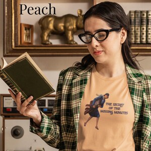 Op de afbeelding: Een perzikkleurig T-shirt met een afbeelding van een persoon in een blauwe jurk die rent met een klok, en de tekst "THE SECRET OF THE MISSING MINUTES" in donkerblauw. Het shirt wordt gedragen onder een groen-wit geruite blazer.