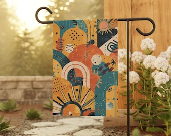 Op de afbeelding: Een kleurrijke tuinvaandel met een abstract ontwerp met zonnen, bogen en geometrische vormen in tinten oranje, geel en teal. De vlag wordt getoond op een zwarte metalen standaard in een tuin.