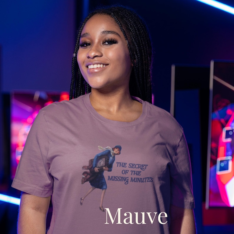 Op de afbeelding: Een mauve t-shirt met korte mouwen en een grafische illustratie van een persoon in een blauwe jas die rent terwijl diegene een klok vasthoudt. De tekst "THE SECRET OF THE MISSING MINUTES" staat in donkerblauwe letters naast de illustratie.