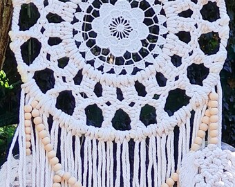 Puede incluir: Un atrapasueños de macramé blanco con un diseño circular, con un patrón floral central y borlas colgantes. Cuentas de madera y plumas decorativas añaden detalles. El atrapasueños está suspendido al aire libre.