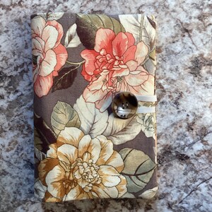 May include: A brown fabric wallet with a floral pattern featuring pink, white, and brown flowers. The wallet has a brown button closure and a white elastic band.
