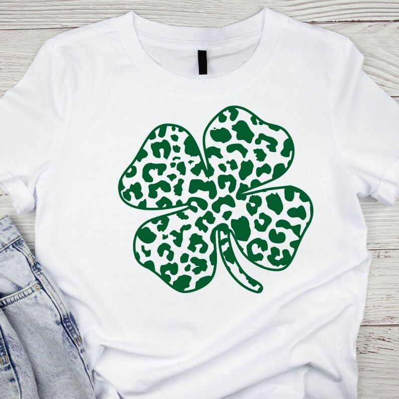 Pode incluir: Camiseta branca com um design de trevo de quatro folhas verde. O trevo &eacute; feito de um padr&atilde;o de estampa de leopardo.